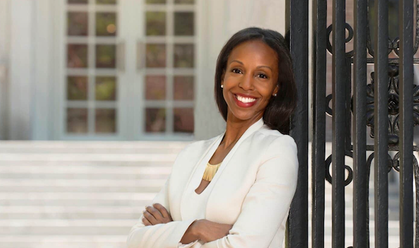 a middle aged black woman smiles at the camera leaning against a rod iron fence. Her arms are crossed and she is wearing a white suite with a cream colored shirt visible underneath. She has medium length dark hair, bright white teeth and is wearing diamond stud earrings.