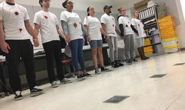 A row of students stand together, holding hands; they wear white t-shirts stained with red marks as a protest against gun violence