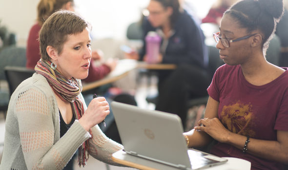 Sociology professor kneeling down and talking to a student who is sitting at a desk in class with an open laptop