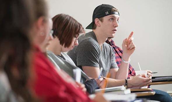 students sitting in desks in a BSU Special Education class including a male student with short light brown hair wearing a gray t-shirt and black baseball cap backwards who is talking and gesturing