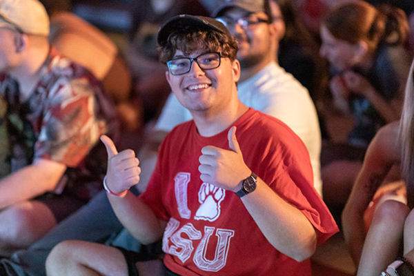 2022.08.31_crimson_celebration_au-20-600x400.jpg A BSU student smiling and giving thumbs up, wearing glasses, a baseball cap and an I paw BSU tshirt, sitting on a floor surrounded by other students