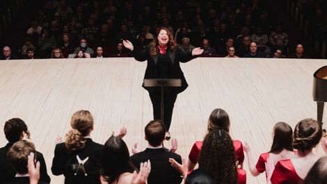 Colleen McDonough opens her arms as she conducts a choir on a stage.
