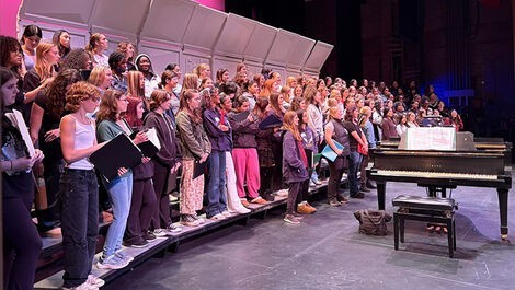 A large group of students sing on a stage next to a piano.