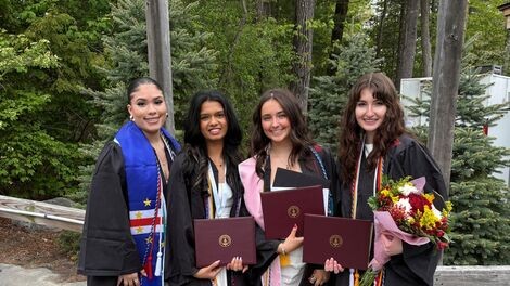 Four female students wearing graduation gowns stand in front of trees.