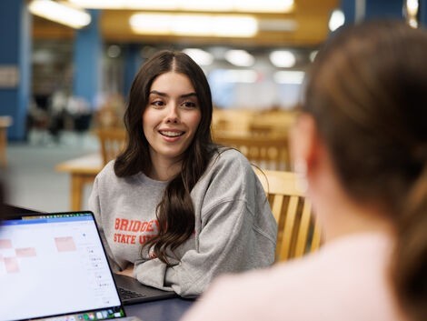 A woman with long brown hair smiles while talking to another woman 