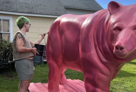 A person stands next to a pink bear and uses a paintbrush to paint