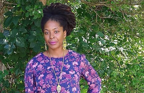 A woman wearing a purple dress stands in front a green tree and poses for camera