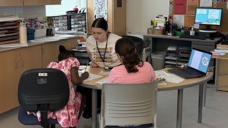A student works with children while all are sitting at a table in a classroom.