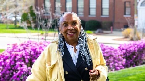 Sheryl Royster stands in front of purple flowers while wearing a yellow jacket.