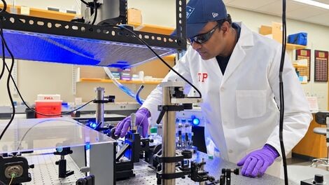 A professor in a white lab coat works at a table filled with optical equipment.