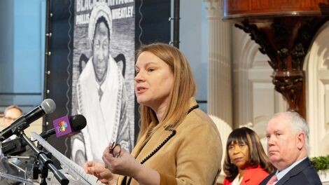 Emily Field speaks from behind a podium with a picture of the Phillis Wheatley stamp behind her.