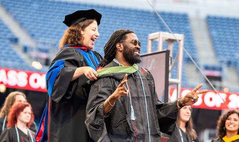 Dr. Lisa Boehm, Dean of the College of Graduate Studies smiles widely as she puts a master's degree hood on a BSU graduate who smiles and makes the peace sign with both hands