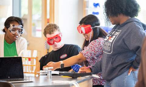 Four students wearing protective goggles collaborating on an experiment in the lab