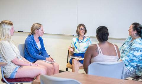 Professor Elissa Pototsky sits in a circle with 4 students in front of the whiteboard in a classroom teaching 