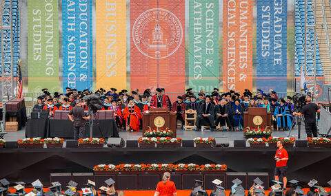 The stage at Commencement 2025 with President Clark speaking at a podium, faculty and board members behind him and rows of graduates in their caps and gowns looking on