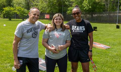3 members of BSU Athletics smiling standing in front of BSU cornhole games