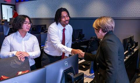 A student shaking hands with Governor Healy in the BSU cyber range. Another student looks on.