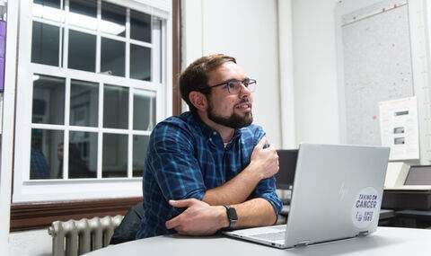a male BSU student with glasses and short dark brown hair, mustache and beard looking up from his laptop