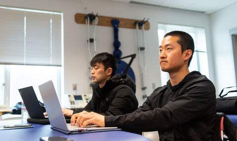 2 graduate students in class sitting at a padded table with laptops open and crutches on the wall in background