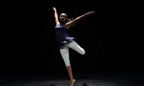 female dancer on stage wearing light gray cropped leggings and a royal purple tunic top standing on one pointed toe with her arms up and outstretched and her left leg bending behind her in a dance