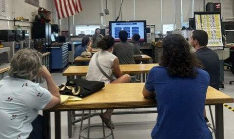 In a vocational shop 4 tables of 2 people each look at a large television screen and listen to a speaker at the front of the room