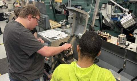 In a machine shop 2 students look at paperwork while working at a machine