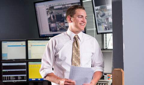 Phillip Rondelli, '17, smiling wearing a white button up shirt with a tan tie. He is holding paperwork with several computer screens in the background
