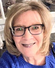 Donna Abelli smiling with medium length dirty blonde hair and wearing black rim glasses and a royal blue sweater