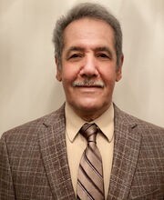 Dr. Jabbar Al-Obaidi smiling with short gray hair and mustache. He is wearing a brown suit jacket over a tan button up shirt and a gold tie with brown stripes