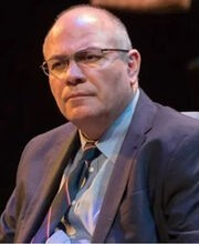 Dr. Francisco Alatorre balding with short gray hair wearing brown rim glasses a light blue button down shirt, navy blue tie and blazer