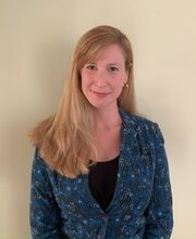 Maria Armour smiling with long dirty blonde hair parted on the side and wearing a black top under a blue, black, white and yellow print blazer