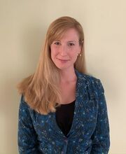 Maria Armour smiling with long dirty blonde hair parted on the side and wearing a black top under a blue, black, white and yellow print blazer