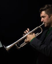 Dr. Spencer Aston with short dark brown hair playing trumpet while looking at sheet music. He is wearing black on a black background