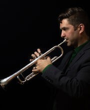 Dr. Spencer Aston with short dark brown hair playing trumpet while looking at sheet music. He is wearing black on a black background