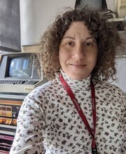 Dr. Margaret Black with medium length brown curly hair wearing a white with print turtleneck and a red lanyard standing in front of old computers
