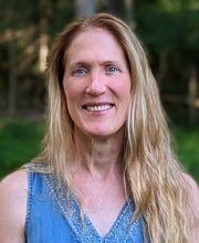 Dr. Darcy Boellstorf smiling with long straight dirty blond hair wearing a v-neck sleeveless blue top with grass and trees in photo background