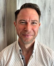 Adam Brieske Ulenski smiling with short brown hair and stubble mustache and beard. He is wearing a white button down shirt with small black print