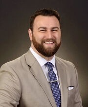 Dr. Lance Brooks smiling with short brown hair, beard and mustache. He is wearing a tan suit jacket over a white button down shirt and blue tie
