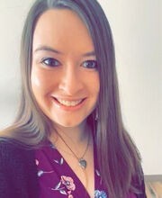 Christine Camara smiling with long straight brown hair and wearing a purple wrap dress with a black sweater