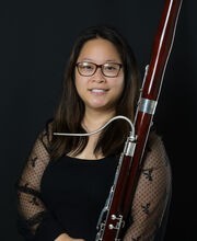 Debbie Chen smiling with long dark brown hair and wearing black rim glasses and a black dress with sheer sleeves. She is holding a bassoon.