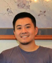 Dr. Xiangfei Chen smiling with short black hair and a short goatee. He is wearing a dark gray t-shirt/jersey with framed artwork behind him