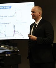 Dr. CJ Cleary teaching next to a projector screen and smiling while wearing a black suit and tie with a tan button down shirt