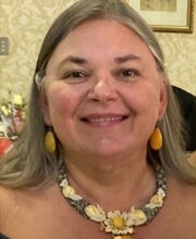 Veronica Cote smiling with medium length gray hair and wearing a black off the shoulder dress with gold colored earrings and necklace