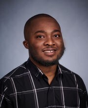 Dr. Edwin Dartey smiling with shaved black hair and a short mustache and beard. He is wearing a black button down shirt with large gray box stripes