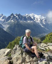 Dr. Susan Eliason sitting on top of a mountain smiling with short gray hair wearing blue shorts, t-shirt, backpack and hiking sneakers