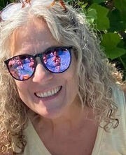 Jan Feldmann smiling with medium length curly gray hair and wearing black rimmed mirrored sunglasses and a sleeveless yellow top