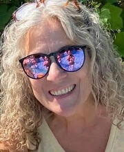 Jan Feldmann smiling with medium length curly gray hair and wearing black rimmed mirrored sunglasses and a sleeveless yellow top
