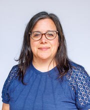 Dr. Susan Flis smiling with medium length straight graying brown hair and wearing brown rimmed cat's eye shaped glasses and a short sleeve blue top