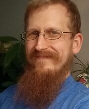 headshot photo of Stephen Flood with short reddish brown hair, mustache and beard wearing metal rimmed glasses and a blue jersey