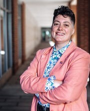 Dr. Reihonna Frost-Calhoun smiling with short dark brown curly hair wearing a salmon colored blazer with rainbow flag pin on it and blue and white print button down shirt under it